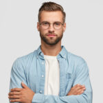 Horizontal shot of handsome self confident businessman or designer, stands crossed hands against white background, wears casual shirt, poses against white background. People and lifestyle concept